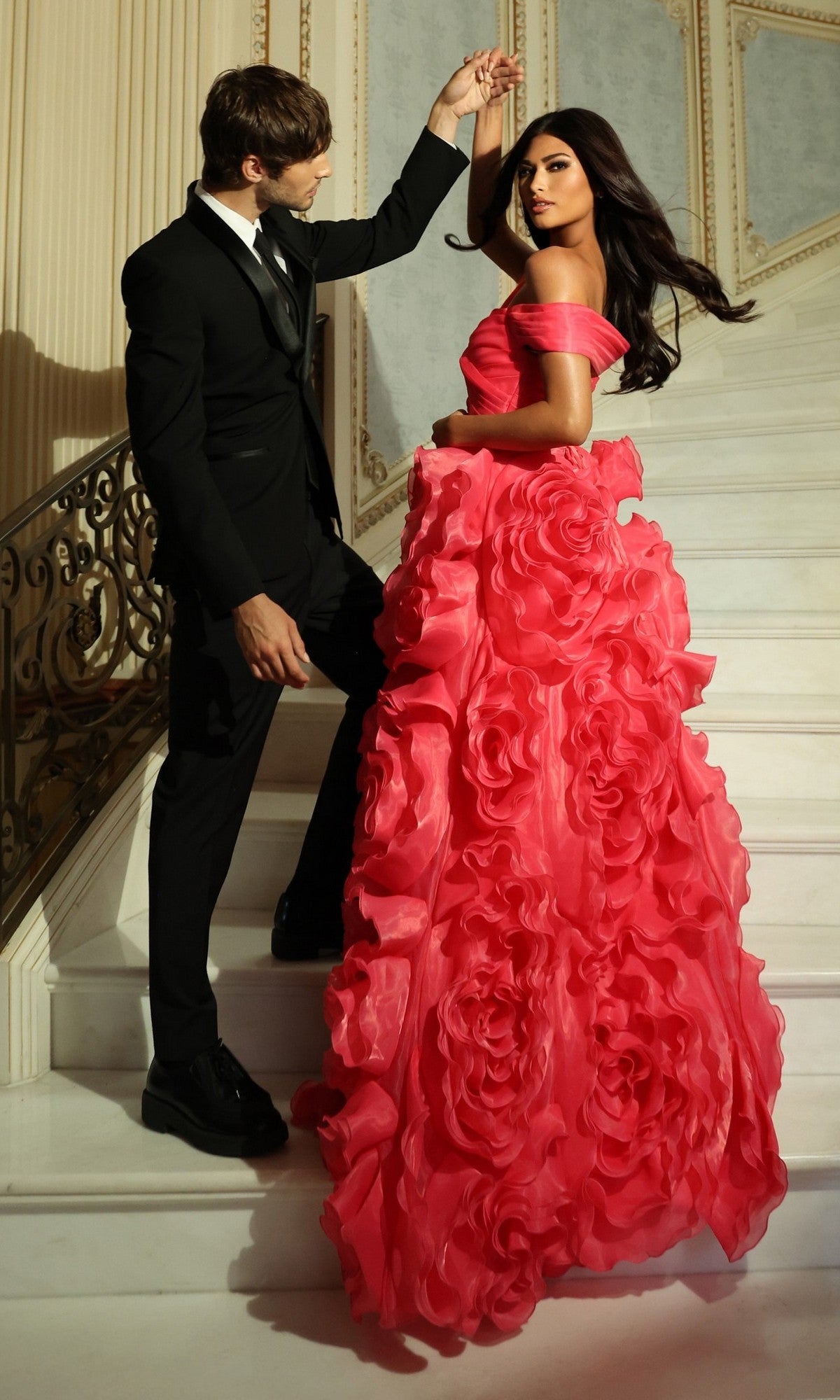 A man in a black suit and a woman in the vibrant Jovani 42187 long prom dress pose together on an elegant staircase.